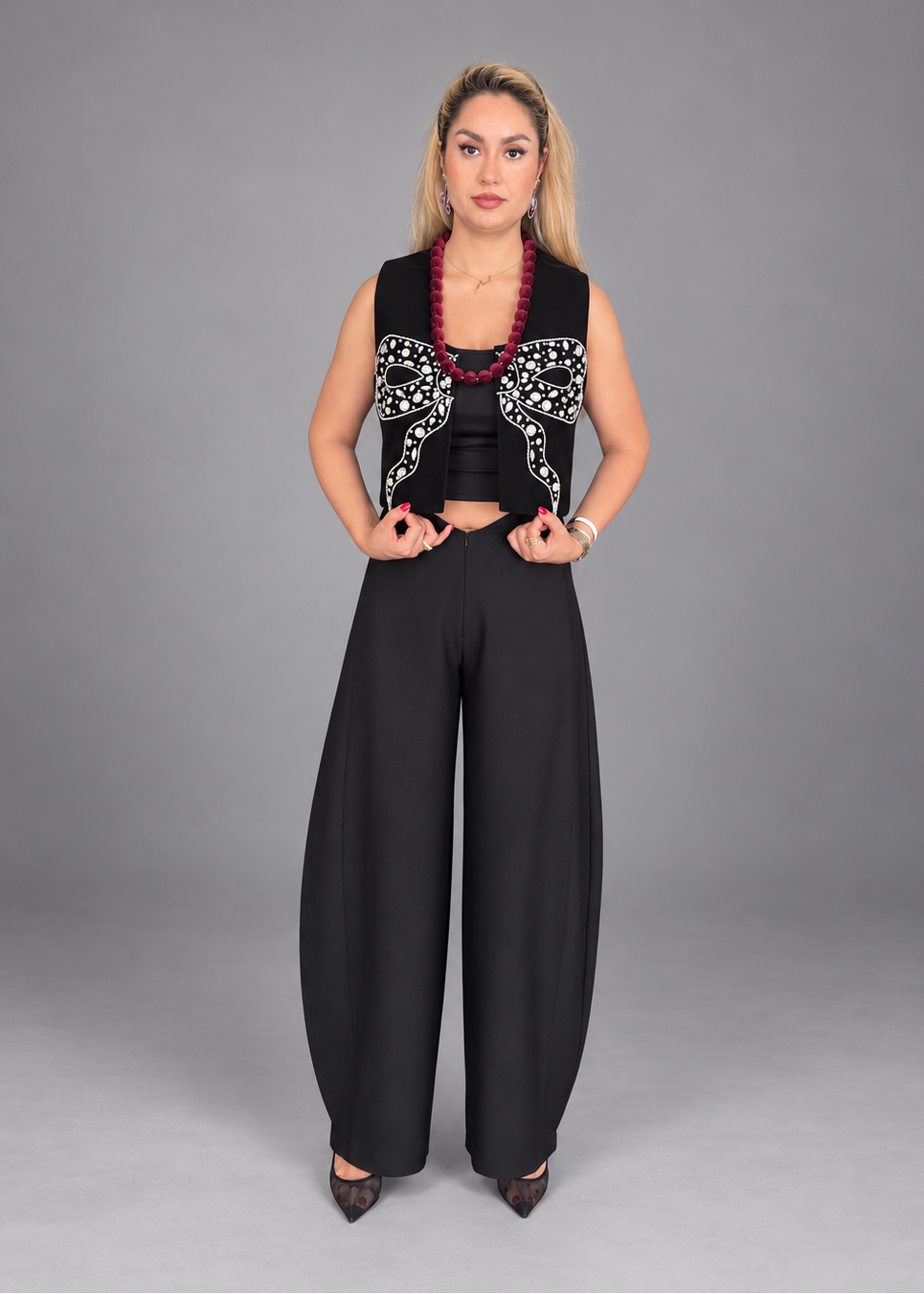 A woman modeling a black sleeveless vest with white embroidered bow details, paired with wide-leg black pants, a black crop top, and a red beaded necklace, against a solid gray studio background.