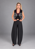 A woman modeling a black sleeveless vest with white embroidered bow details, paired with wide-leg black pants, a black crop top, and a red beaded necklace, against a solid gray studio background.
