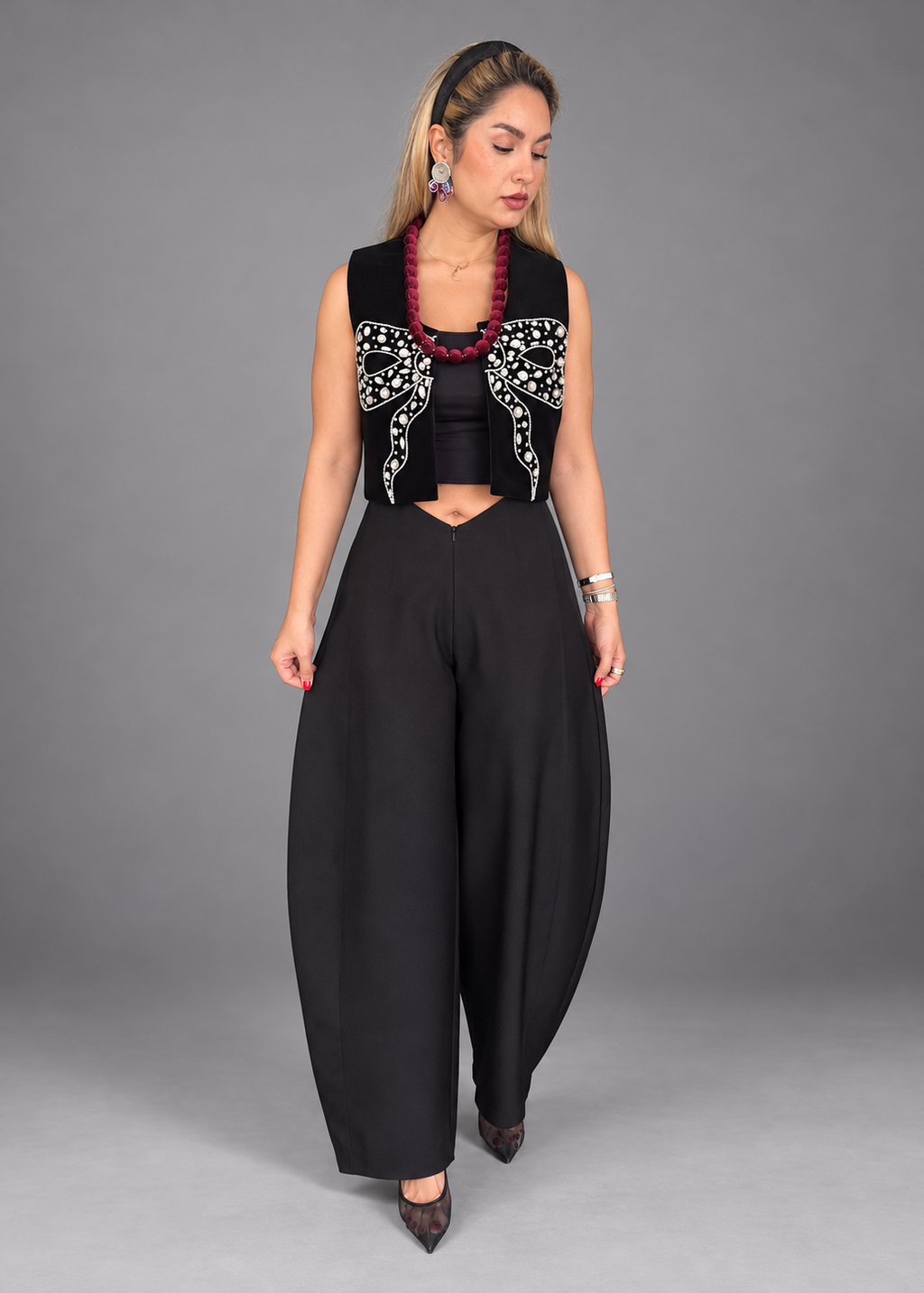 A woman models a sleeveless black vest with intricate silver embroidery on the chest, paired with high-waisted black wide-leg pants and a red beaded necklace, against a solid gray studio background.
