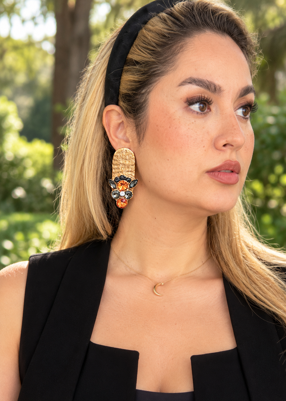 A woman wearing a black headband and black sleeveless top, featuring a large statement earring with a gold-toned textured top and a colorful floral drop in orange, blue, and black, set against a blurred green outdoor background.