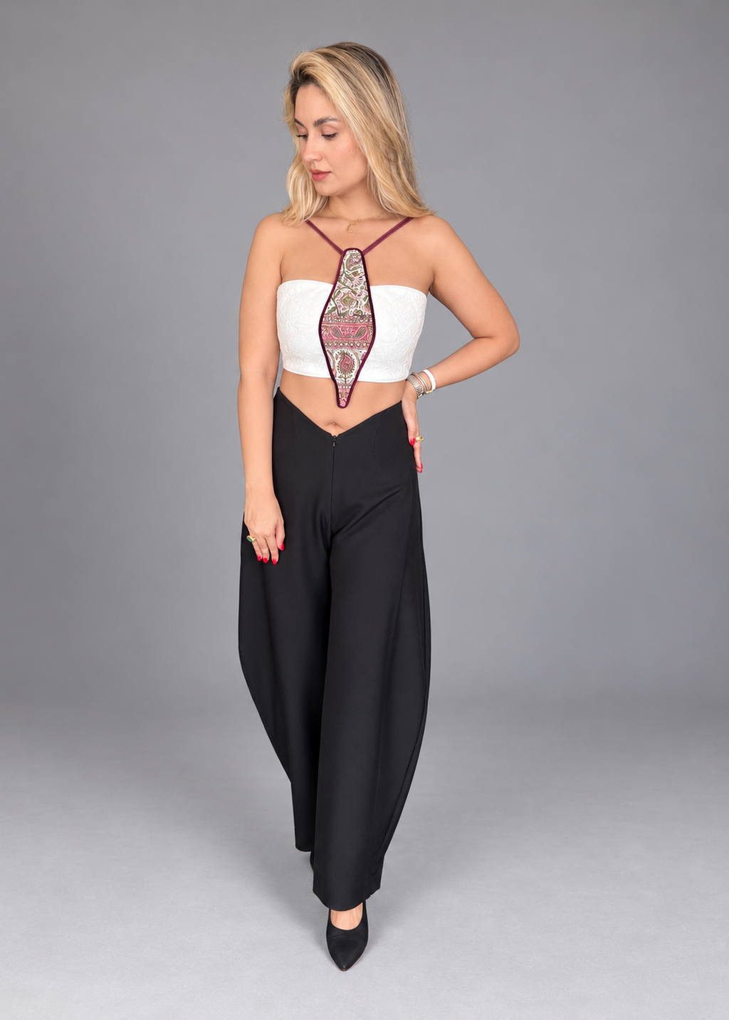 A woman modeling a two-piece outfit consisting of a white strapless crop top with a burgundy and gold embroidered tie detail, paired with high-waisted black wide-leg pants, against a solid gray studio background.