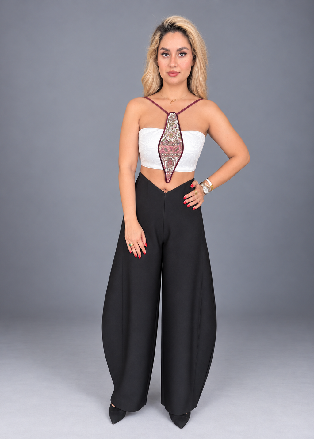 A woman modeling a two-piece outfit consisting of a white strapless crop top with a central embroidered panel and black wide-leg pants, shot against a solid gray studio background.