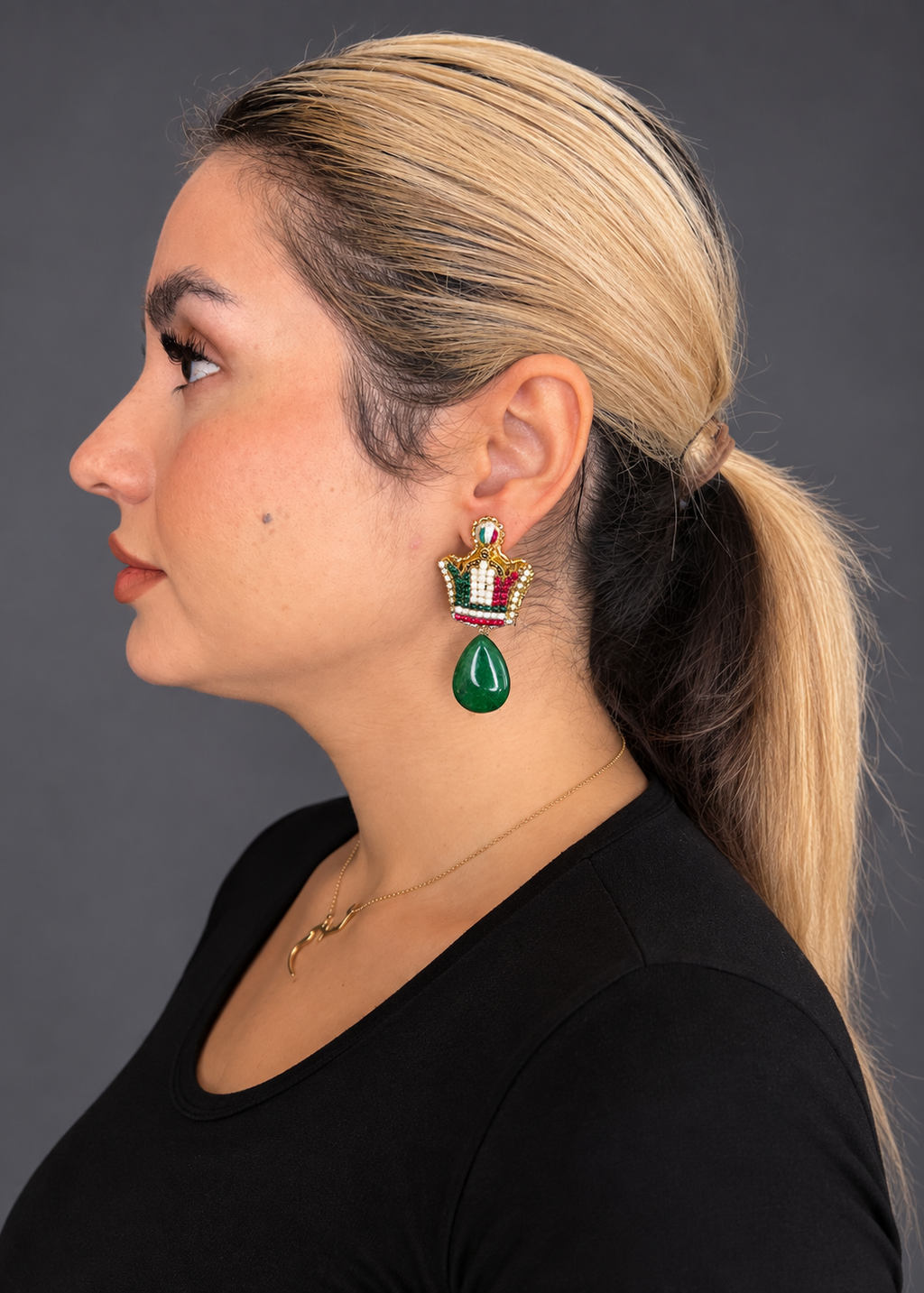 A woman in profile wearing a gold crown-shaped drop earring with a green teardrop pendant and red and white enamel details, against a dark gray studio background.