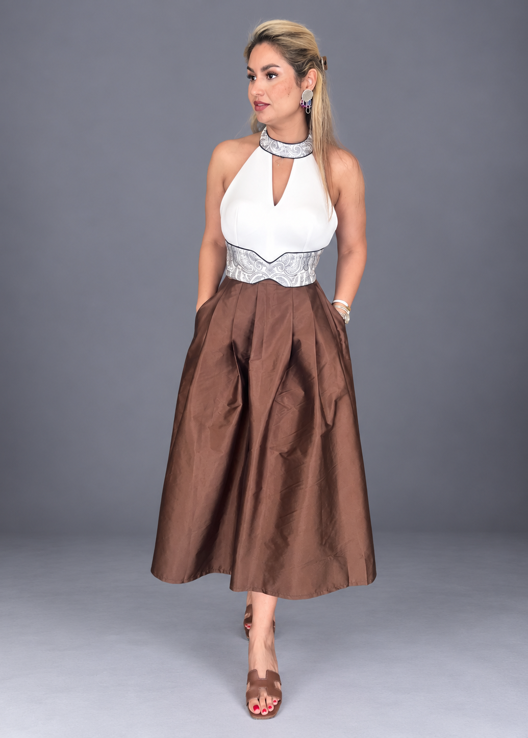 A woman models a formal midi dress featuring a white halter top with a keyhole cutout and silver patterned waistband, paired with a voluminous brown pleated skirt, set against a solid gray studio background.