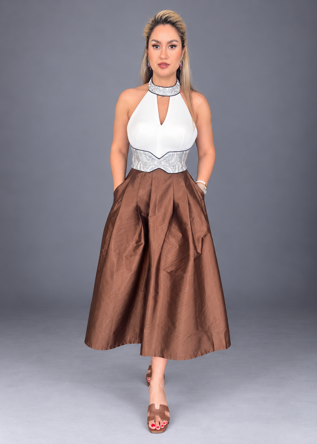 A woman modeling a sleeveless midi dress with a white halter-style top, silver embellished waistband, and a voluminous brown skirt, posed against a solid gray studio background.