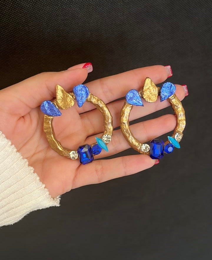 Gold hoop earrings with blue and clear gemstones held in a hand against a black background