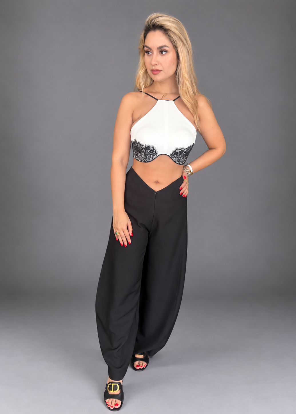 A woman modeling a two-piece outfit consisting of a white halter crop top with black lace trim and black wide-leg pants, standing against a solid gray studio background.