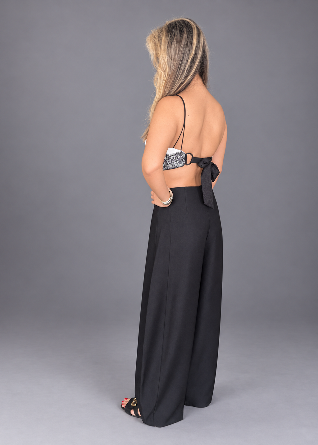 A woman shown from the back wearing a black wide-leg jumpsuit with thin straps and a bow detail at the waist, paired with black strappy heels, against a solid gray studio background.