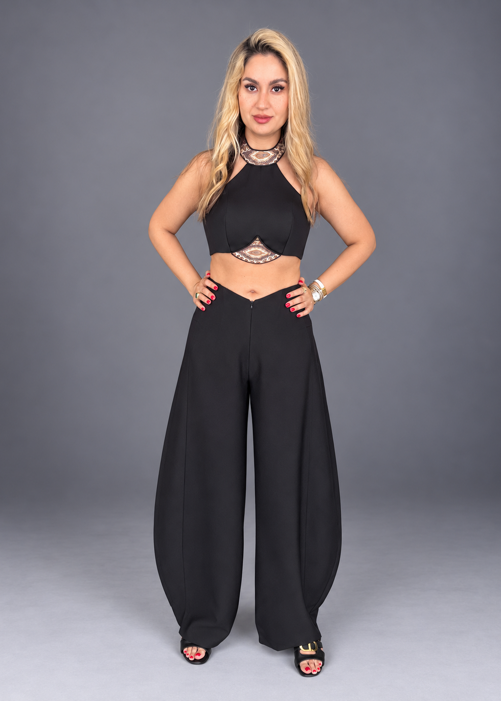 A woman modeling a black two-piece outfit consisting of a halter crop top with a decorative cutout and matching high-waisted wide-leg pants, posed against a solid gray studio background.