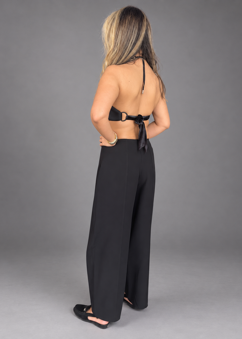 A woman modeling a black two-piece outfit consisting of a halter crop top with lace cutouts and matching wide-leg pants, photographed against a solid gray studio background.