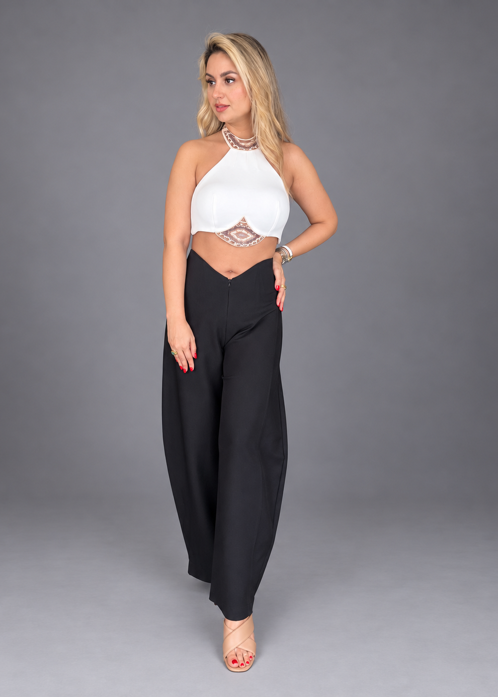 A woman modeling a white halter crop top with a lace waist detail and black wide-leg pants against a solid gray studio background.