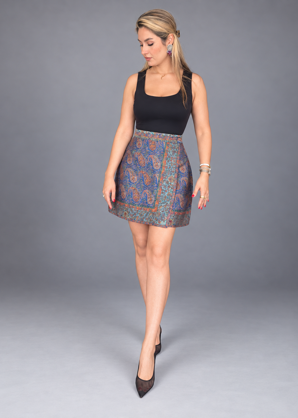 A woman modeling a sleeveless black top and a high-waisted mini skirt featuring a blue and orange paisley pattern, posed against a solid gray studio background.