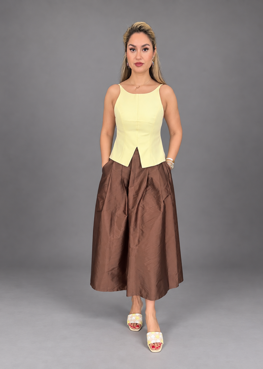 A woman modeling a sleeveless two-piece outfit consisting of a pale yellow corset-style top and a brown wide-leg midi skirt, paired with white slide sandals, against a solid gray studio background.