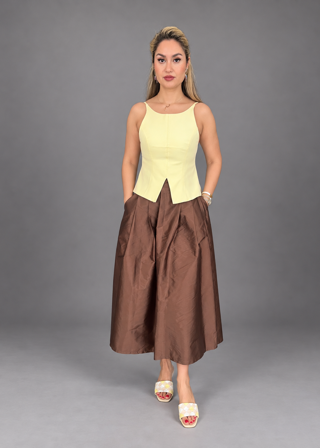A woman modeling a sleeveless two-piece outfit consisting of a pale yellow corset-style top and a brown wide-leg midi skirt, paired with white slide sandals, against a solid gray studio background.