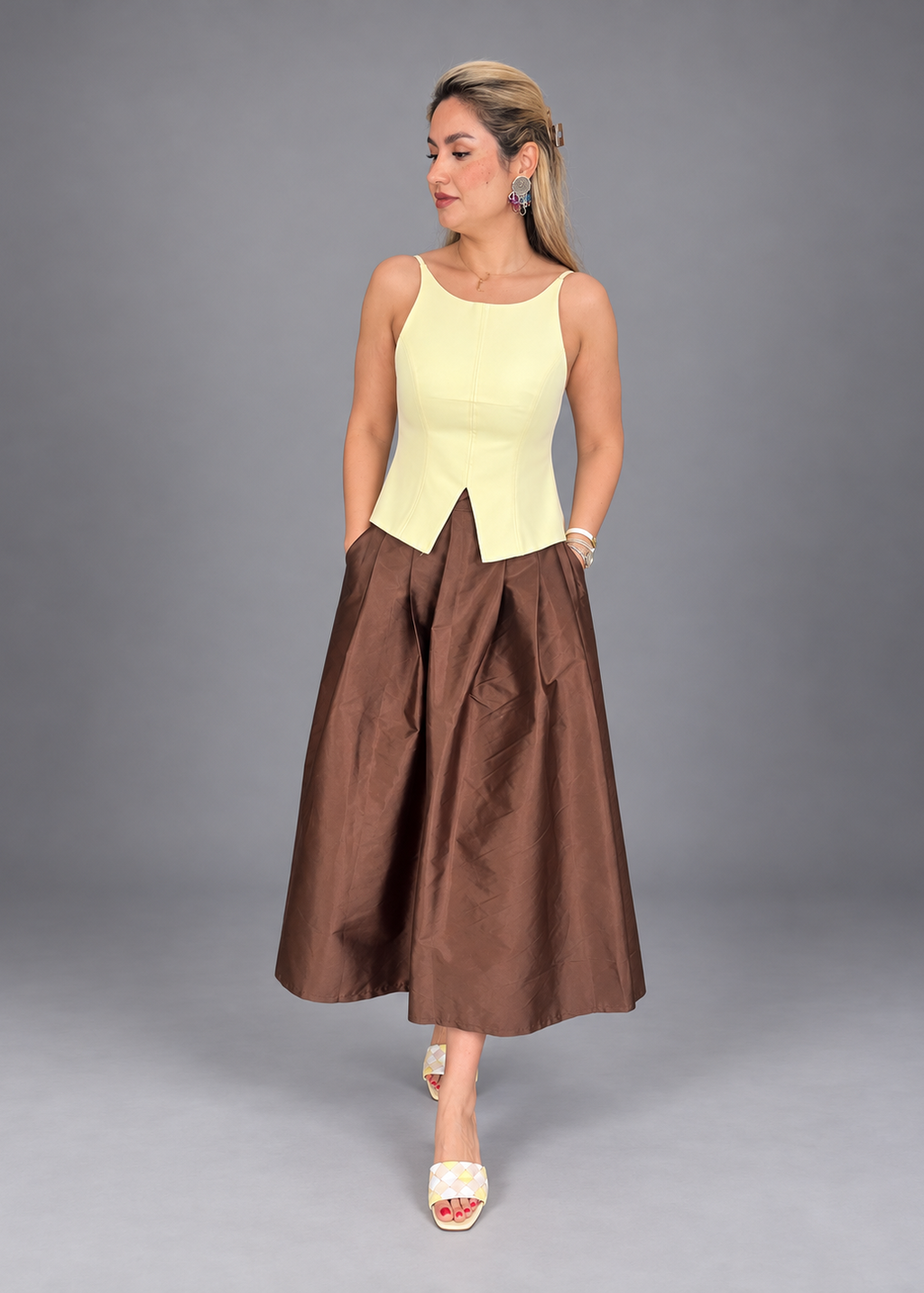 A woman models a sleeveless midi dress featuring a pale yellow structured bodice and a voluminous brown skirt, posed against a solid gray studio background.