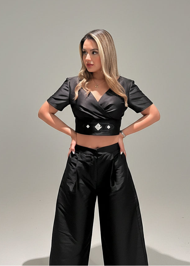 Woman wearing a black outfit with a crop top and wide-leg pants against a gray background