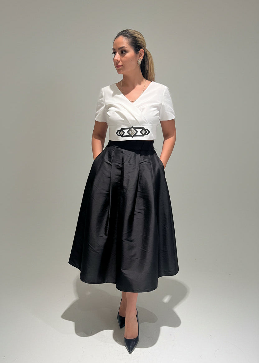Woman wearing a white top with a black belt and black skirt against a beige wall.