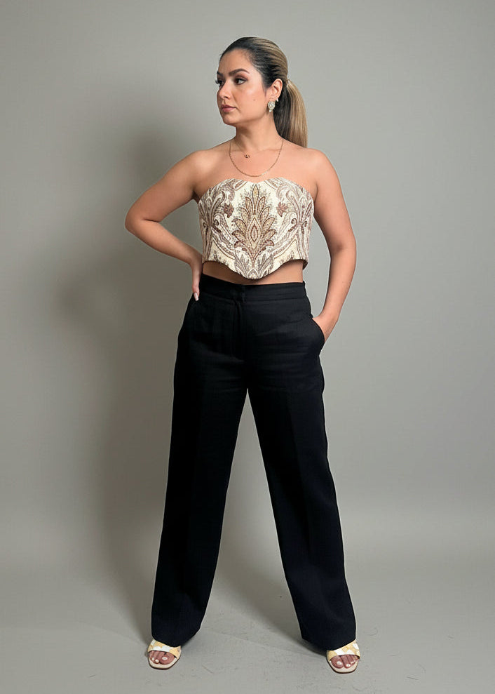 Woman wearing a strapless top with intricate design and black pants against a gray background