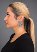 A woman in profile wearing a black top and a gold necklace, showcasing a colorful, multi-component drop earring with a silver-toned circular medallion and dangling red, blue, green, and white elements, set against a solid gray background.