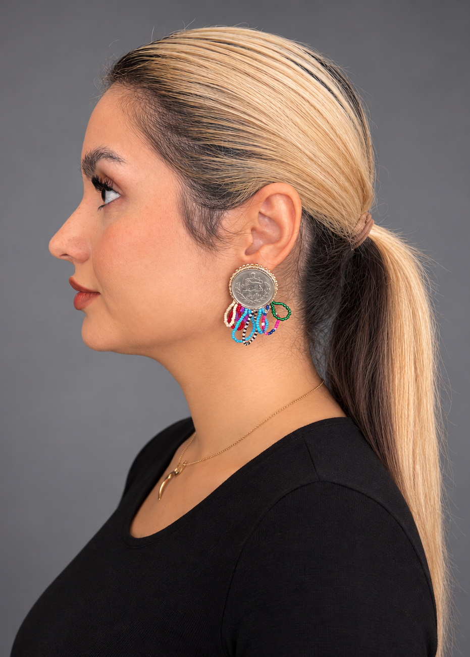 A woman in profile wearing a black top and a gold necklace, showcasing a colorful, multi-component drop earring with a silver-toned circular medallion and dangling red, blue, green, and white elements, set against a solid gray background.