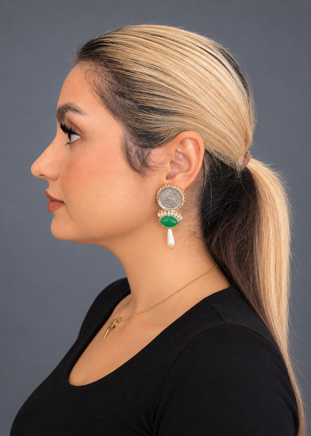 A woman in profile wearing a gold-toned earring with a silver-toned circular disc, a green bead, and a white dangling tassel, against a dark gray background.