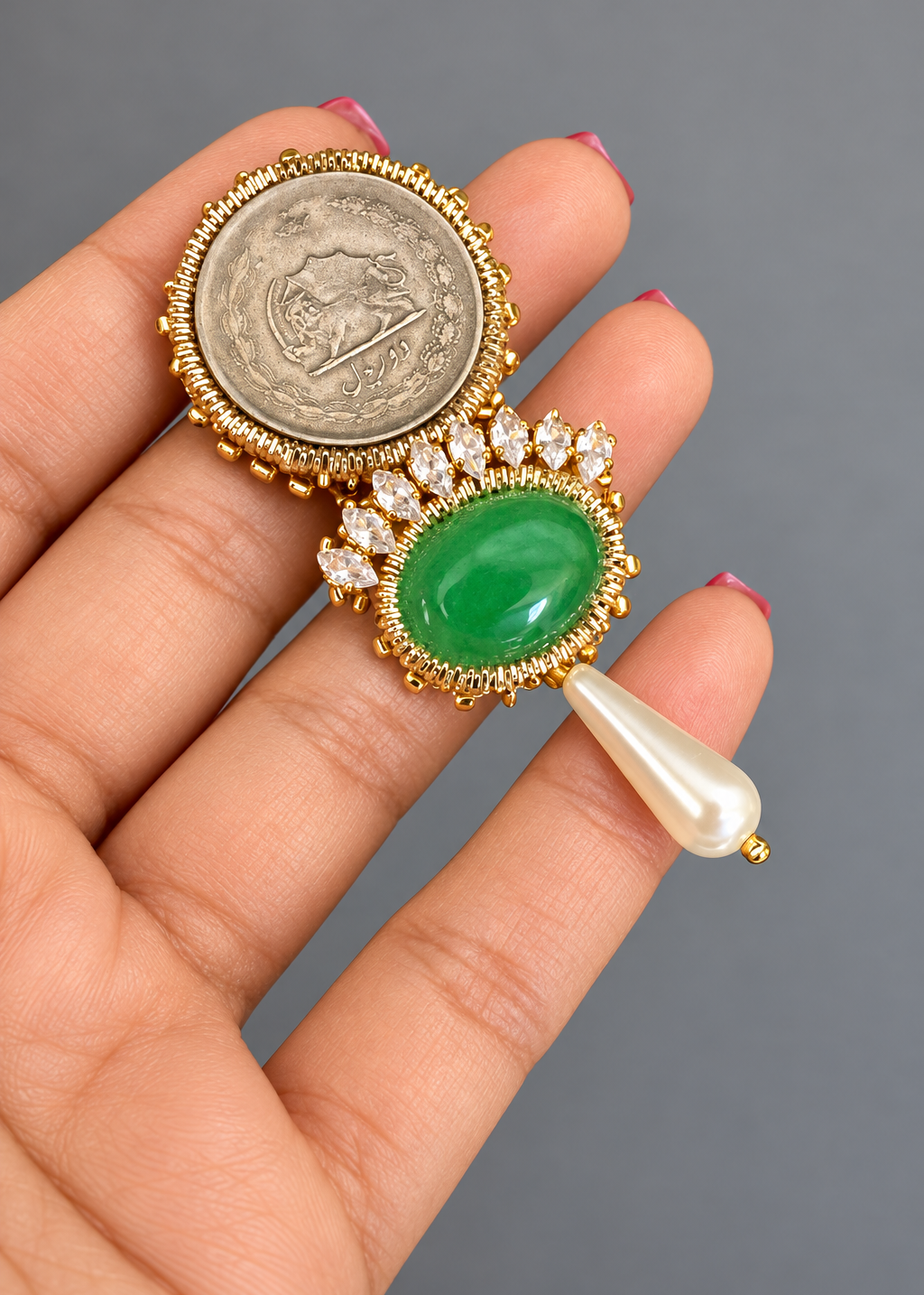 A gold-toned jewelry piece displayed on a person's hand, featuring a round silver coin, a green oval stone surrounded by small clear stones, and a dangling white pearl-like drop.
