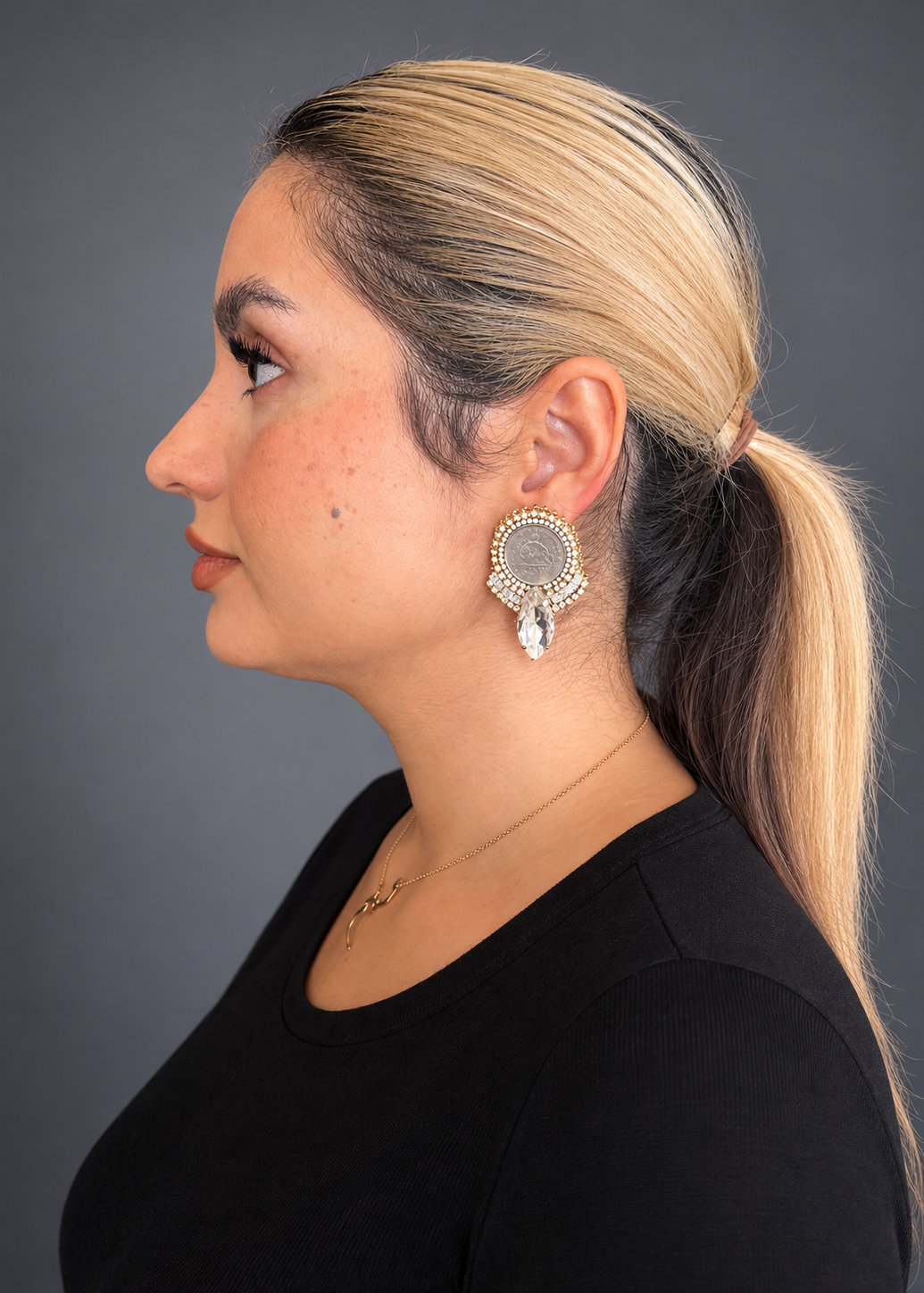 A woman in profile wearing a large silver-toned circular earring with a dangling clear crystal, shown against a dark gray studio background.