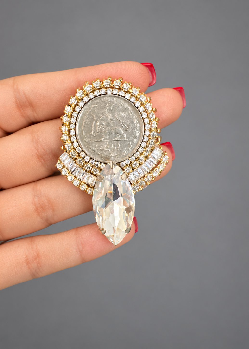 A hand with red-painted nails holds a gold and silver coin brooch featuring a large marquise-cut clear crystal pendant, set against a solid gray background.