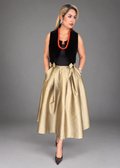 A woman models a sleeveless black velvet vest over a black top, paired with a voluminous champagne-gold satin midi skirt featuring gold floral accents at the waist, and black pointed-toe heels, against a solid gray studio background.