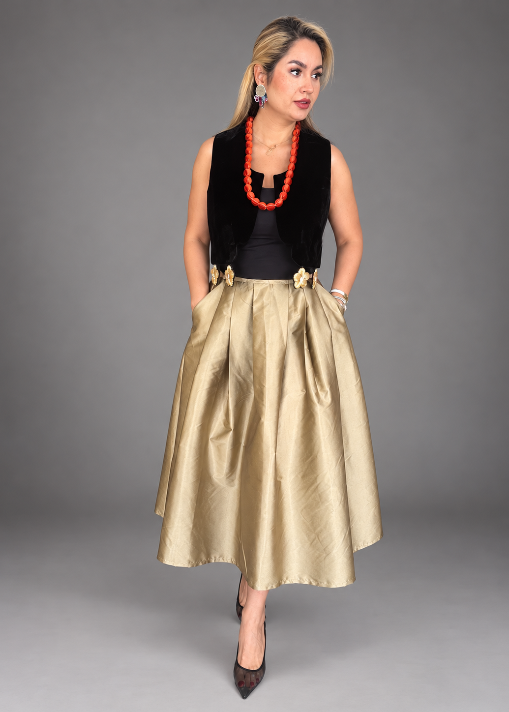 A woman models a sleeveless black velvet vest over a black top, paired with a voluminous champagne-gold satin midi skirt featuring gold floral accents at the waist, and black pointed-toe heels, against a solid gray studio background.