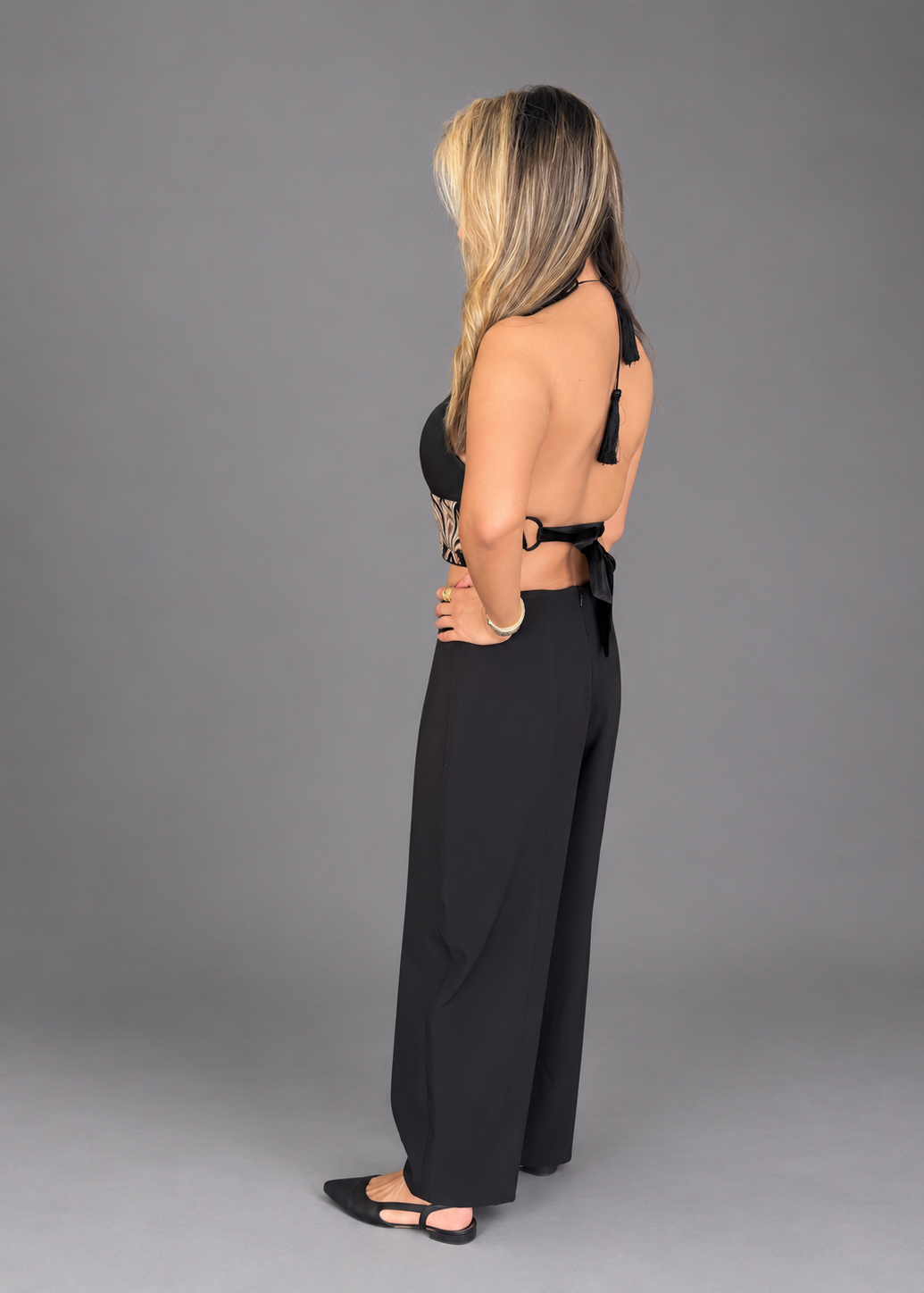 A woman modeling a black two-piece outfit consisting of a sleeveless crop top with gold floral embroidery and a matching high-waisted wide-leg pants, set against a solid gray studio background.