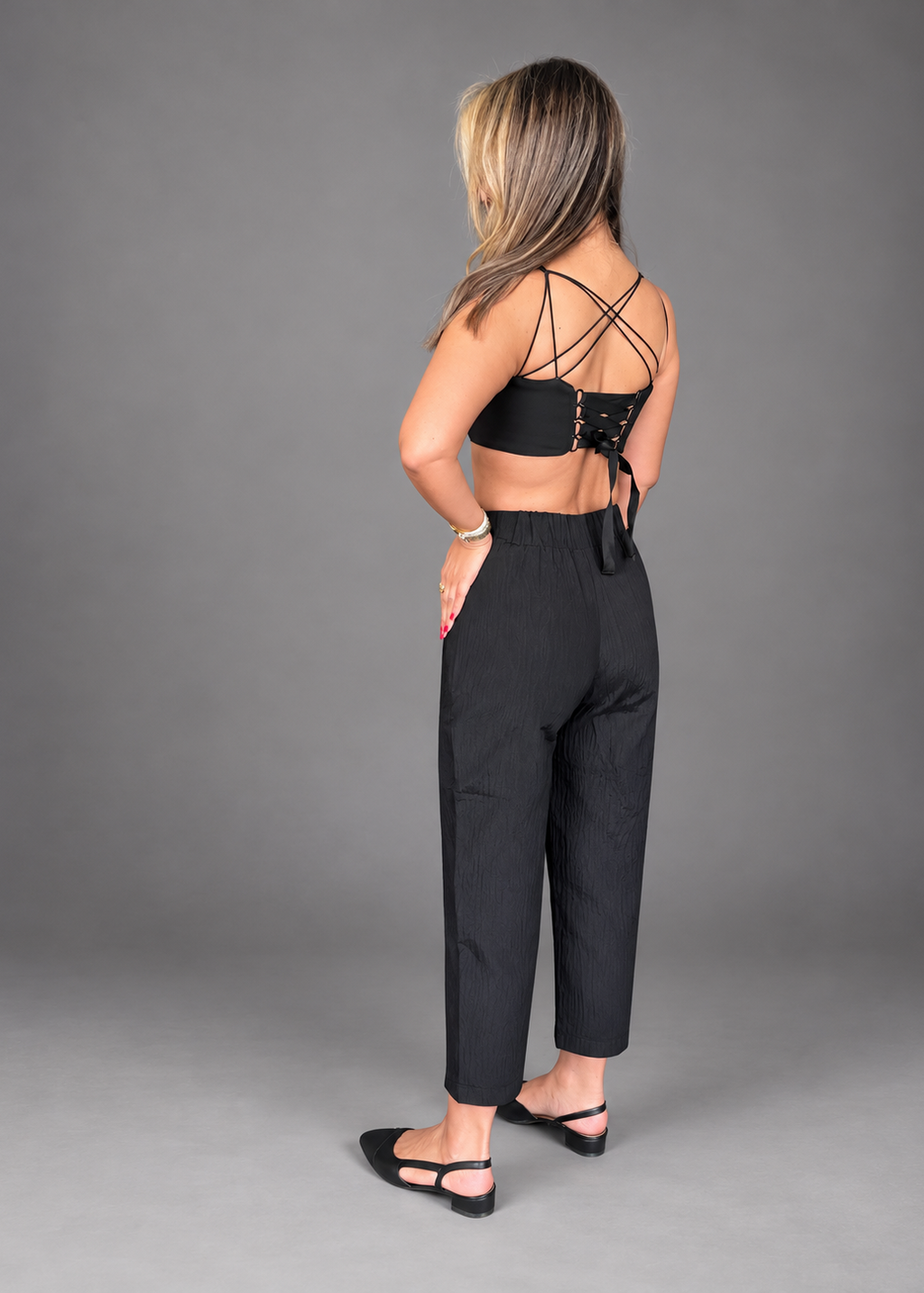 A woman is shown from the back wearing a black strappy lace-up crop top and black cropped trousers, standing against a solid gray studio background.