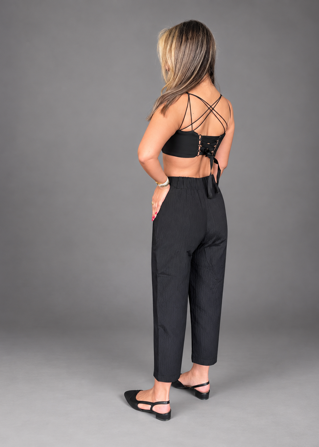 A woman is shown from the back wearing a black strappy lace-up crop top and black cropped trousers, standing against a solid gray studio background.