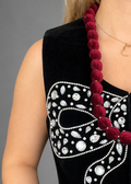 A sleeveless black velvet top with a large white rhinestone bow design on the front, worn by a model with a dark red beaded necklace.