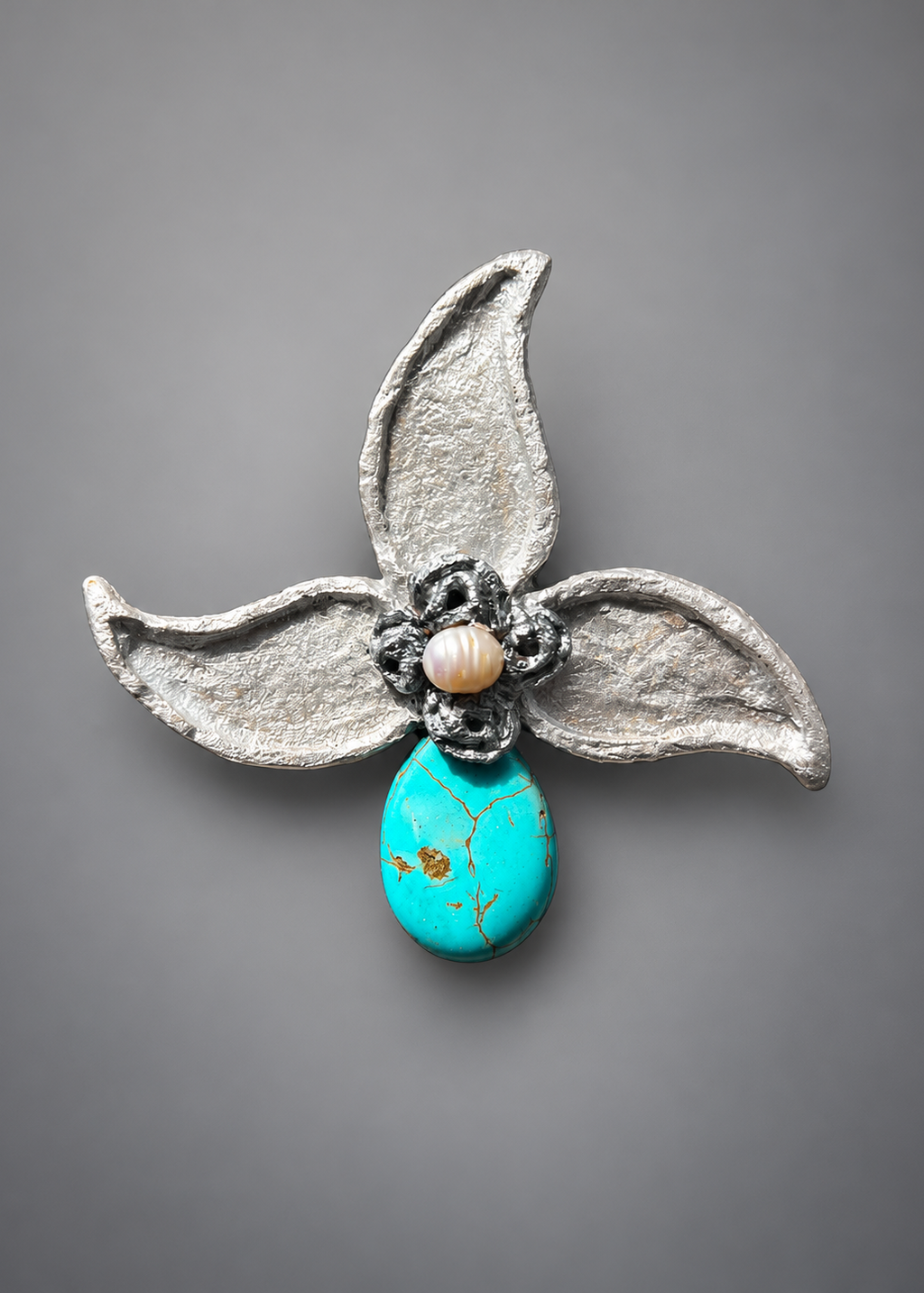 A handmade flower-shaped brooch featuring textured silver petals, a dark gray and white beaded center with a small cream pearl, and a turquoise teardrop pendant with gold flecks, set against a solid gray background.