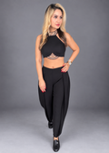 A woman modeling a black two-piece outfit consisting of a halter crop top with lace detailing and matching high-waisted wide-leg pants, set against a solid gray studio background.