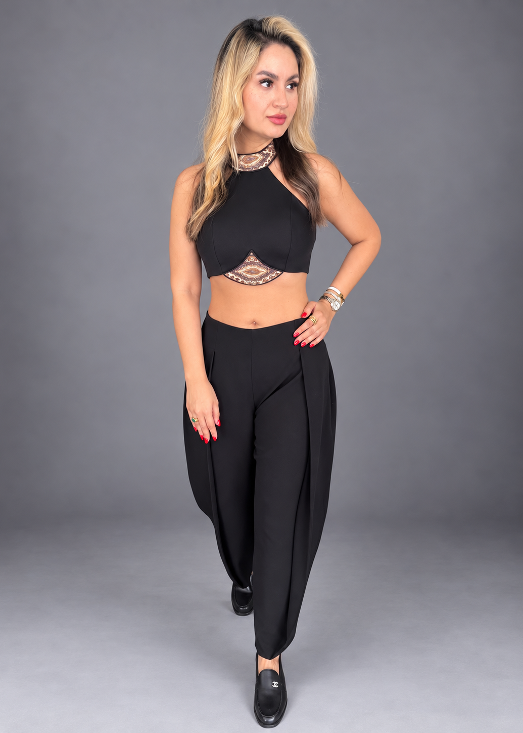 A woman modeling a black two-piece outfit consisting of a halter crop top with lace detailing and matching high-waisted wide-leg pants, set against a solid gray studio background.